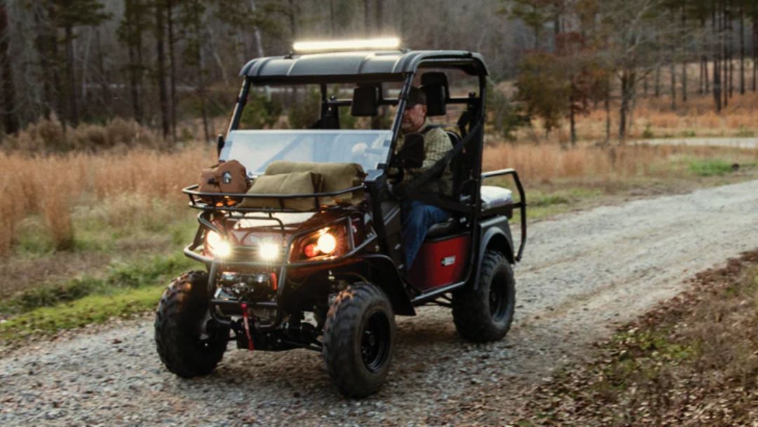 Bad Boy Buggy utility vehicle with headlights on driving over bumpy gravel road, illustrating conditions that cause front-end suspension noise