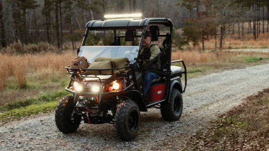 Bad Boy Buggy utility vehicle with headlights on driving over bumpy gravel road, illustrating conditions that cause front-end suspension noise